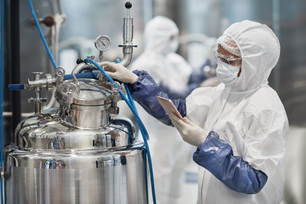 Zwei Personen in weißen Schutzanzügen mit Kapuzen, Gesichtsmasken und blauen Gummihandschuhen arbeiten an einem großen, glänzenden Kessel in einer industriellen Anlage. Die Person im Vordergrund bedient ein Tablet.